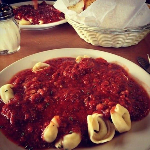 Plates of tortellini with red sauce, a cheese shaker, and a bread basket on a wooden table.